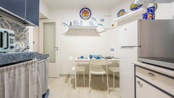 A compact kitchen with a microwave, stove, and refrigerator. There is a dining table with chairs and a small shelf above it.