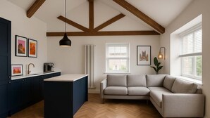 A modern kitchen with dark cabinetry, a central island, and a built-in oven. There is a grey sofa and a window with blinds in the living area. - The Bridge - Penthouse Apartment (York)