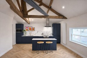 A modern kitchen with dark cabinets, a marble countertop, wooden beams, and a large window. - The Bridge - Penthouse Apartment (York)