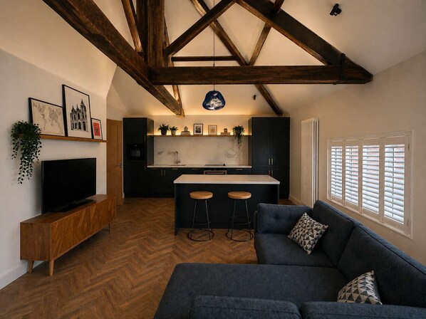 A modern kitchen with dark cabinetry, a central island, and a dining area with a dark sofa and bar stools. - The Bridge - Penthouse Apartment (York)