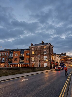 A street view with a building featuring multiple windows and a corner entrance, adjacent to a busier street with vehicles and a cyclist. - The Bridge - Penthouse Apartment (York)