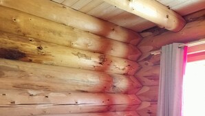 A wooden cabin room with a bed, pillows, a nightstand, a lamp, and a window with curtains.