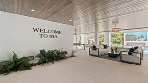 Een moderne lobby met een 'WELCOME TO 81A' bord, een zithoek met banken en een salontafel, en een uitzicht op een zwembad door grote ramen.