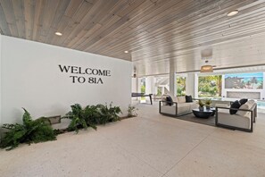 Eine moderne Lobby mit einem Schild mit der Aufschrift 'WELCOME TO 81A', eine Sitzecke mit Sofas und einem Couchtisch sowie ein Blick auf einen Pool durch große Fenster.