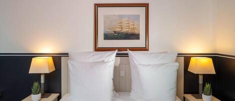 A neatly made bed with white linens, two bedside lamps, and a framed picture of a sailboat on the wall.