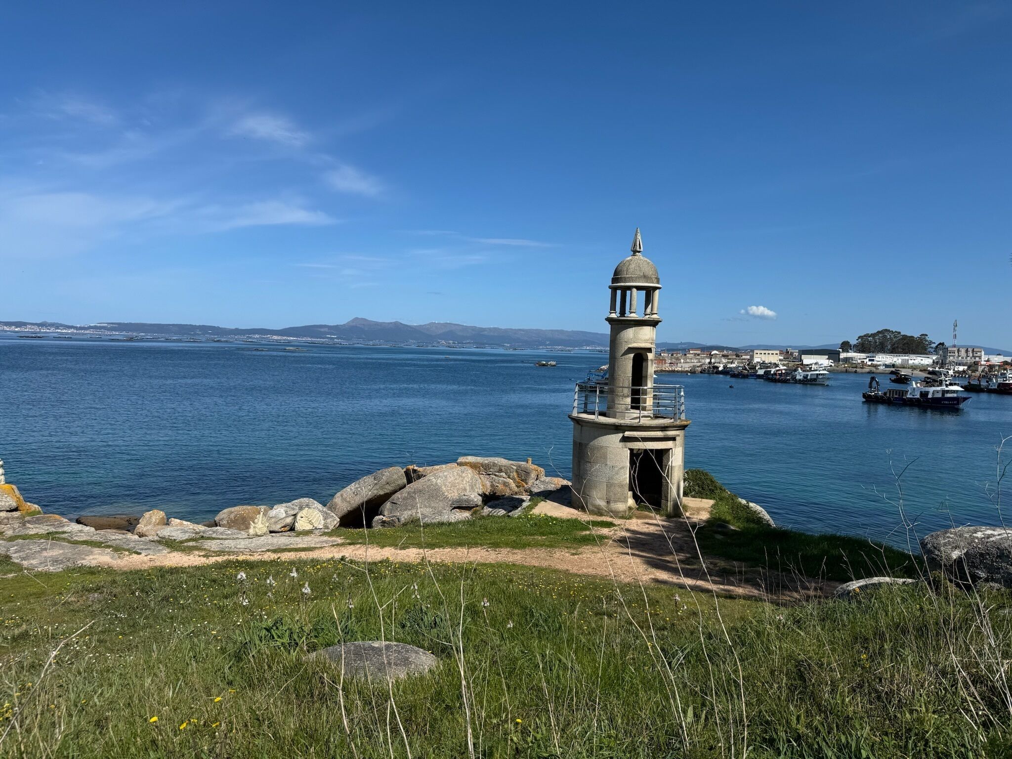 Un faro su una scogliera erbosa che domina un porto con imbarcazioni e edifici sullo sfondo.