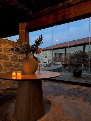 A stone wall, a wooden table with a vase of flowers, and a view of a building with a red roof through large windows. - Luxury room in charming B&B in Galicia (O Agro)