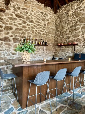 A rustic stone wall bar with a wooden counter, metal bar stools, and shelves with bottles and jars. - Luxury room in charming B&B in Galicia (O Agro)