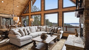A spacious living room with a large sectional sofa, a wooden coffee table, and a stone fireplace. Large windows offer views of the outdoors. - Epic Mountain Views: Hot Tub, Games and Firepit (Ellijay)