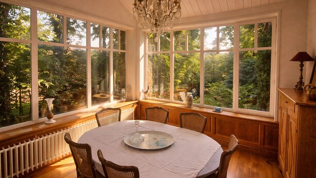 Comedor con una mesa redonda, sillas y una araña. Amplias ventanas ofrecen vistas al exterior con vegetación.