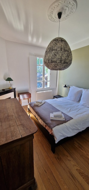 A bedroom with a wooden bed, a hanging lamp, a radiator, a window, and a wooden table.