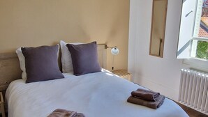 Un dormitorio con cama de madera, ropa de cama blanca, dos almohadas, lámpara colgante, espejo y ventana con luz natural.