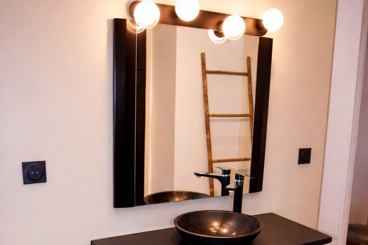 Un meuble-lavabo de salle de bain avec un évier noir, un miroir avec éclairage intégré et une échelle en bois en arrière-plan.