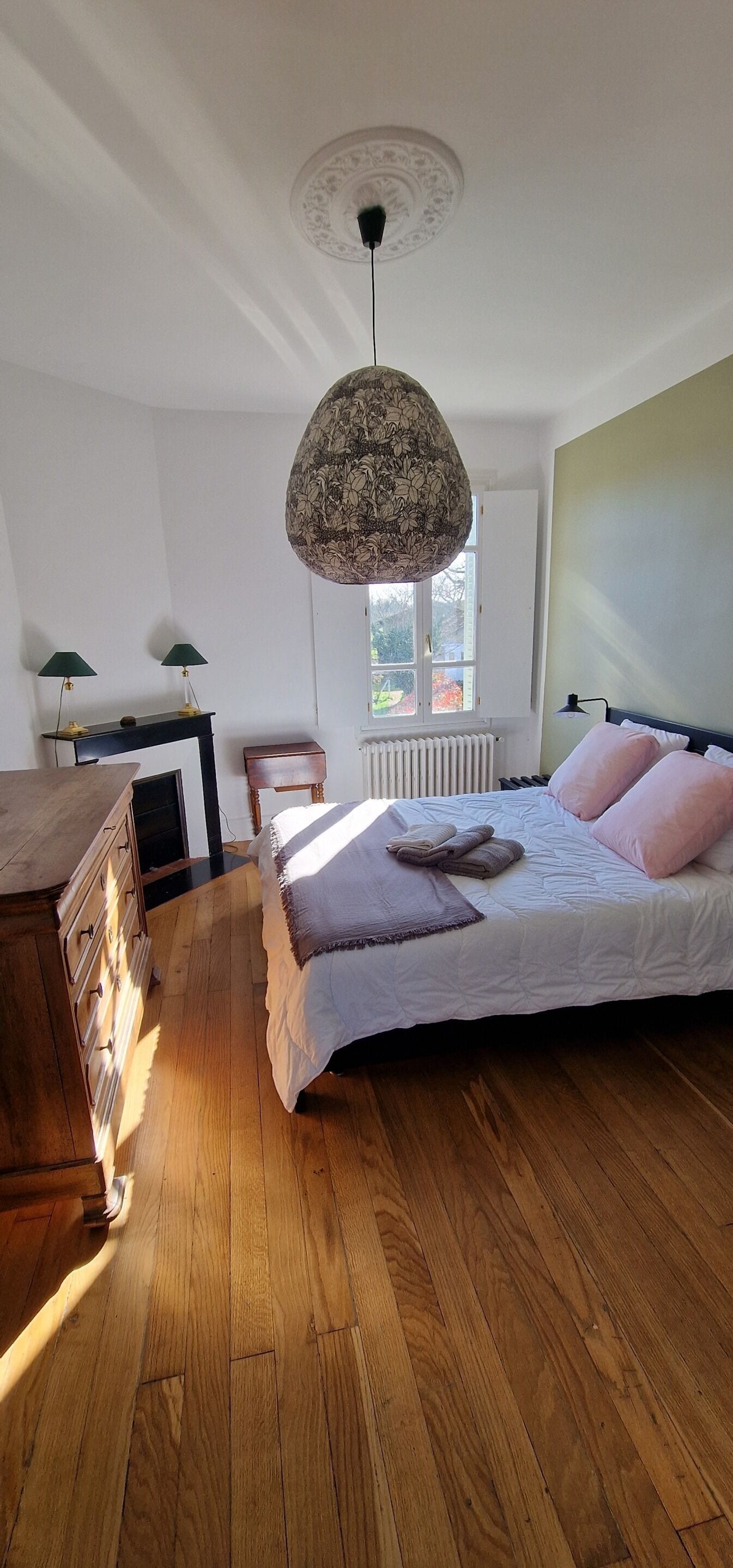 A bedroom with a bed, wooden floor, a desk with lamps, a radiator, and a window.