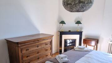 Een slaapkamer met een houten dressoir, een bed met een witte deken, een open haard en een hanglamp.