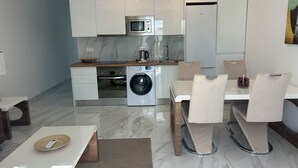A modern kitchen with white cabinets, a built-in oven, microwave, and washing machine. There is a dining table with chairs and a small table with a remote control. - Sunning 2 bedrooom apartment 5 minutes walk to seafront. (Tías)