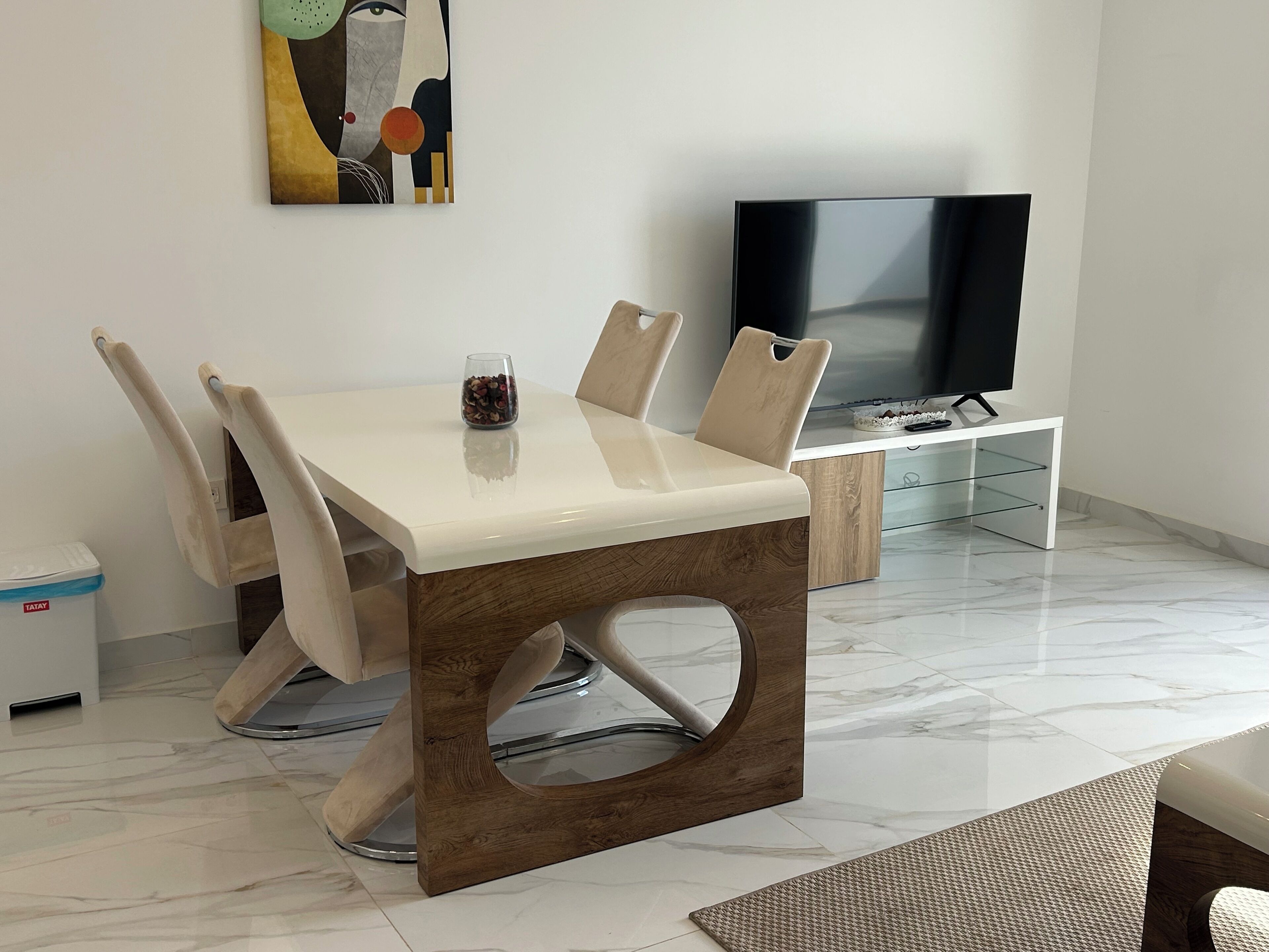 Un espace repas moderne avec une table blanche et des chaises en bois, un vase en verre avec des fleurs, un téléviseur et un meuble blanc.