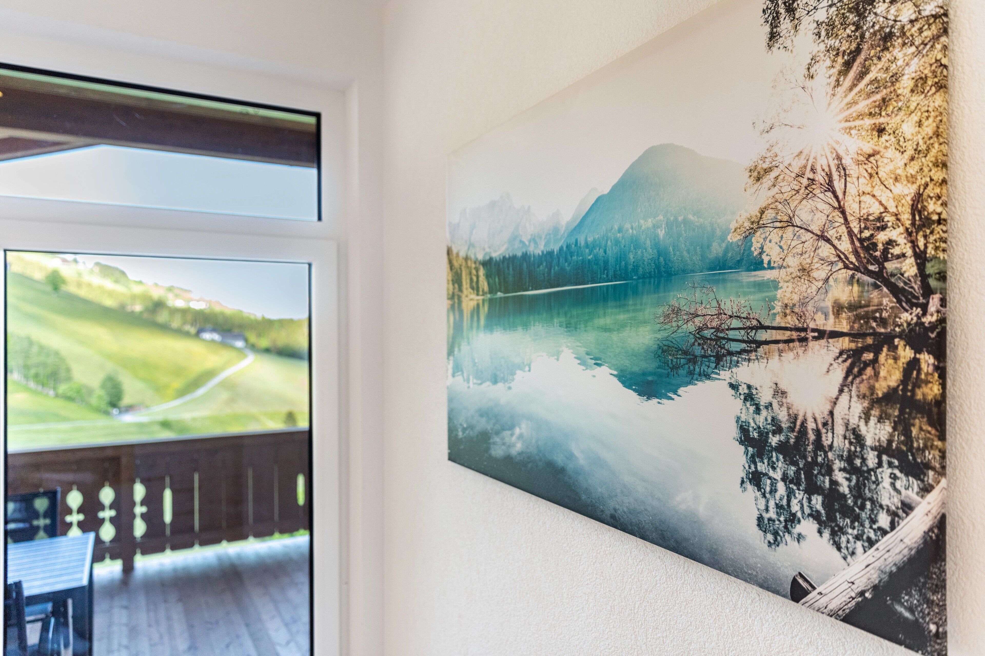 Een kamer met een groot raam dat uitkijkt op een schilderachtig landschap, een houten terras en een afbeelding van een meer en bergen in een lijst.