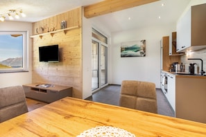 A modern kitchen with a built-in stove, a flat-screen TV mounted on a wooden wall, and a dining area with a wooden table and chairs.