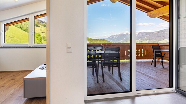 A room with a sliding glass door leading to an outdoor deck with a table and chairs, a wooden ceiling, and a view of a mountainous landscape.