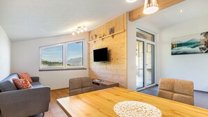 A modern living room with a wooden dining table, grey sofa, and a flat-screen TV mounted on a wooden wall.