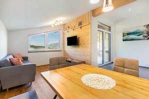A modern living room with a wooden dining table, grey sofa, and a flat-screen TV mounted on a wooden wall.