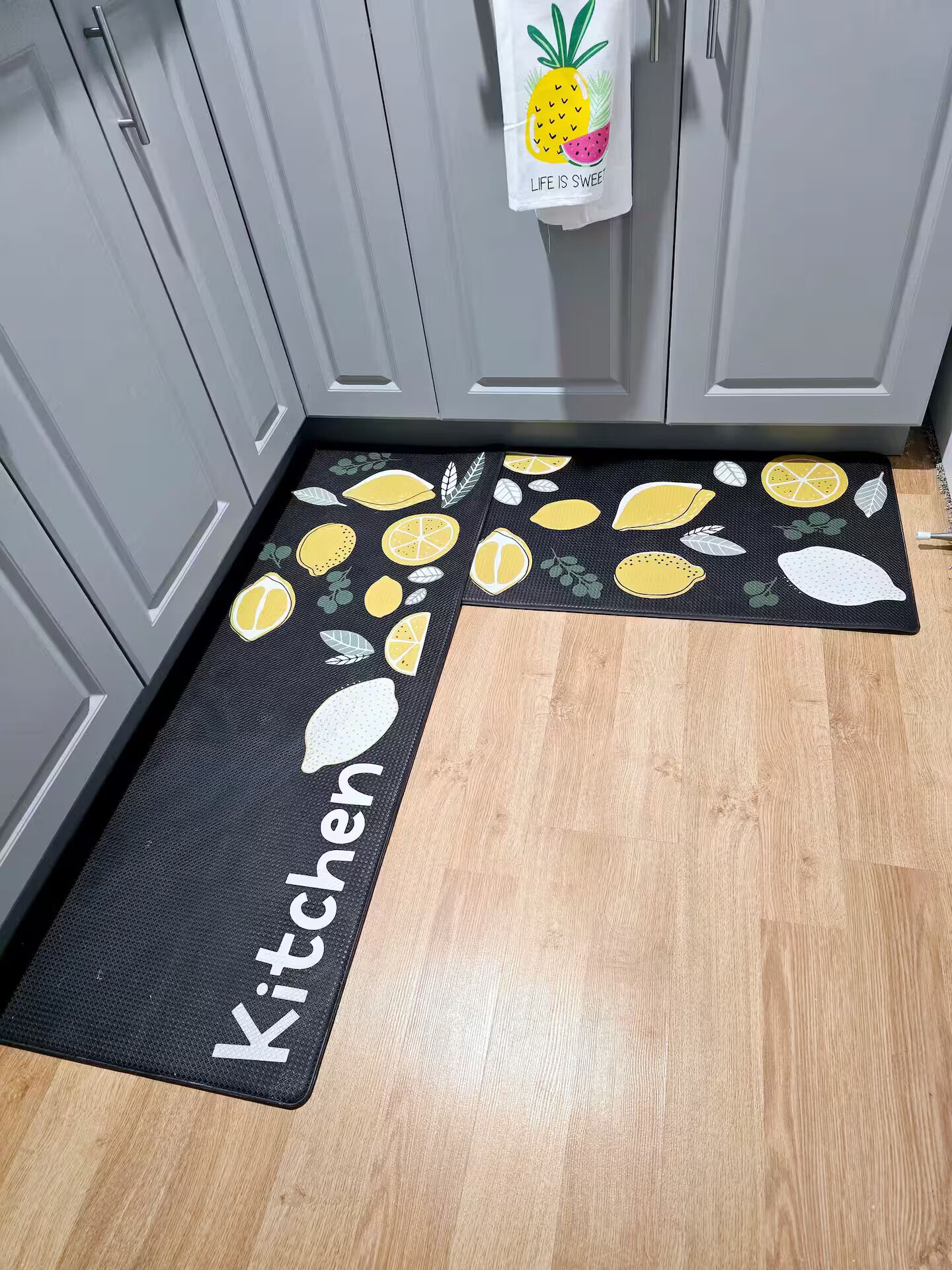 Una cocina con armarios grises, un tapete negro con motivos de limón y piña, y una toalla colgada en la puerta.