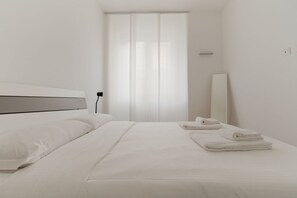Quarto de hotel moderno com uma cama grande, uma mesa de cabeceira, um espelho e uma janela com cortinas leves.