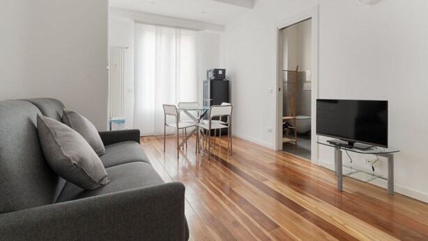 Ein modernes Wohnzimmer mit grauem Sofa, einem Glastisch, einem Flachbildfernseher und einem Essbereich mit weißem Tisch und Stühlen.