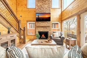 A cozy cabin interior with a fireplace, wooden walls, a staircase, and a large window overlooking a deck. - *Stylish 5BD Lodge w/ VIEWS! | Hot Tub + POOL* (Sevierville)