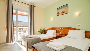 Ein Hotelzimmer mit zwei Betten, einem Balkon mit Vorhängen, einer Wandleuchte und einem Gemälde mit Strandmotiv.