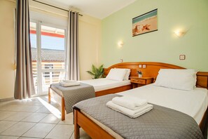 A hotel room with two beds, a balcony with curtains, a wall-mounted light, and a beach-themed painting. - Solena Studios Kavos (Corfu)
