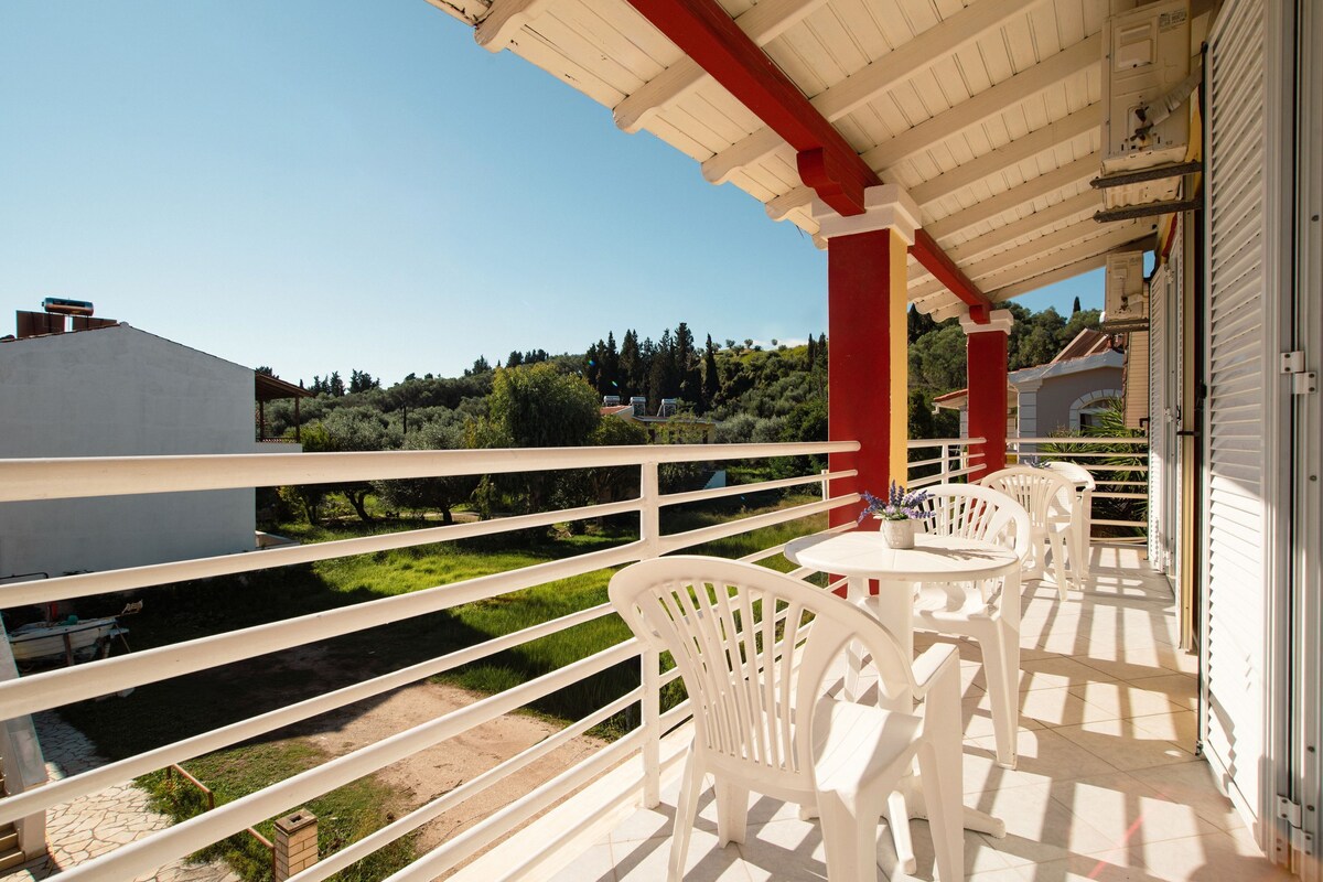 Ein Balkon mit weißen Stühlen und einem Tisch, mit Blick auf eine grüne Landschaft mit Bäumen und Gebäuden.