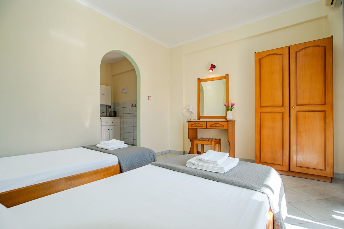 Ein Hotelzimmer mit zwei Betten, einem Holzschrank, einem Schminktisch mit Spiegel und einer kleinen Küchenzeile, die durch einen Bogen sichtbar ist.