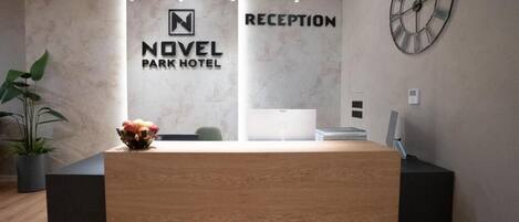 A reception desk at the Novel Park Hotel with a modern design, a clock on the wall, and a potted plant to the left.