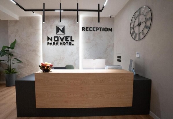 A reception desk at the Novel Park Hotel with a modern design, a clock on the wall, and a potted plant to the left.