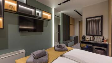 A modern hotel room with a bed, a small desk with a mirror, a television, and a wall-mounted shelf with decorative items.