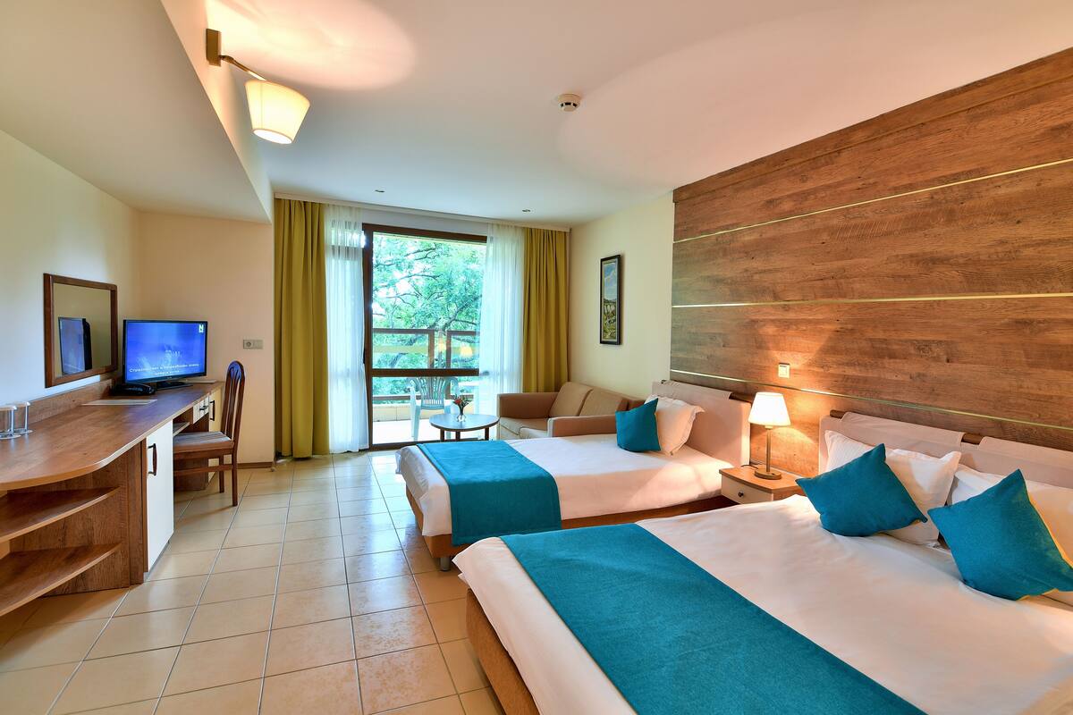 A hotel room with two beds, a desk with a TV, a small table, and a balcony with a view of trees.