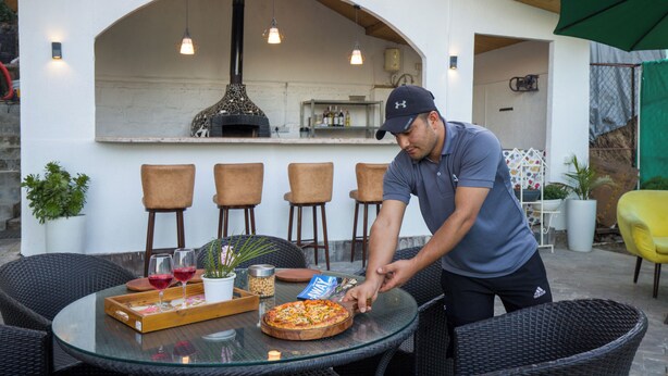 A man cutting a pizza on a glass table with chairs around it, a brick oven in the background, and a bar area. - Alaya Stays 2 BHK with Garden & Hill View | Livelife (Kasauli)