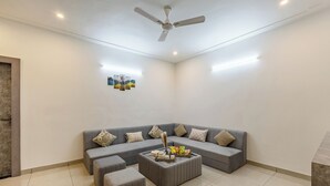 A modern living room with a grey sectional sofa, a grey ottoman, a ceiling fan, and a small table with snacks. - Alaya Stays with Balcony & Hill View | Lake City Heights (Udaipur)