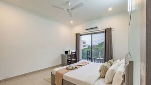 A bedroom with a bed, a desk, and a chair. There is a window with curtains and a view of buildings outside. - Alaya Stays with Balcony & Hill View | Lake City Heights (Udaipur)