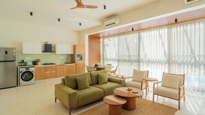 A modern living room with a sofa, chairs, a coffee table, a ceiling fan, and a kitchen area with cabinets and appliances. - ELIVAAS Mirai Leo | 3 BHK Villa Near Vagator Beach With Pool (Vagator)