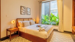 A hotel room with a large bed, a bedside table with a lamp, a window with curtains, and a view of greenery outside. - ELIVAAS Mirai Leo | 3 BHK Villa Near Vagator Beach With Pool (Vagator)