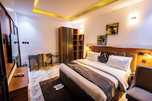 A hotel room with a bed, a desk, a chair, a wardrobe, and artwork on the wall. - Atlantis Hotels & Apartment (Port Harcourt)