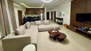 A modern living room with a sectional sofa, a coffee table, a TV, and a kitchen in the background. - LUMAANA RESIDENCES (Abuja)