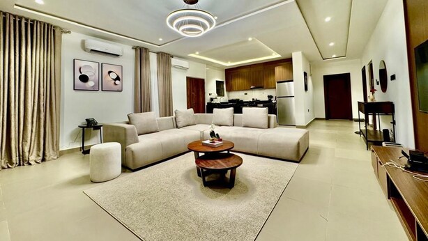 A modern living room with a sectional sofa, a coffee table, and a dining area with a kitchen in the background. - LUMAANA RESIDENCES (Abuja)