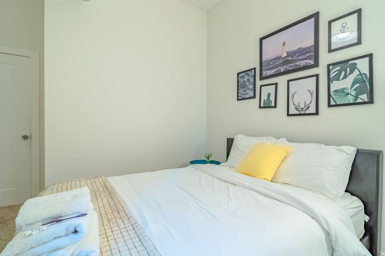 A neatly made bed with white linens and a yellow pillow, a small bedside table with a teal lamp, and a wall adorned with framed artwork including a seascape and botanical prints.