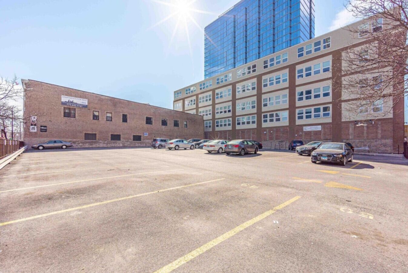A parking lot with several cars, a brick building, and a modern glass building in the background.