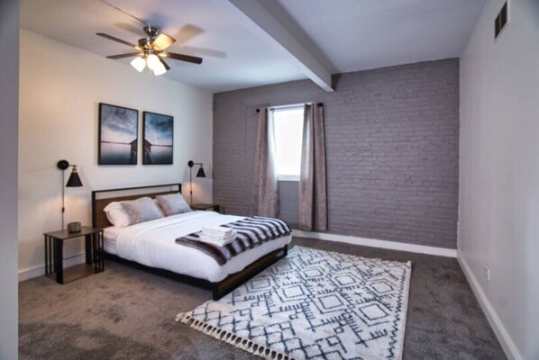 Un dormitorio con cama, ventilador de techo, ventana con cortinas y pared de ladrillo.