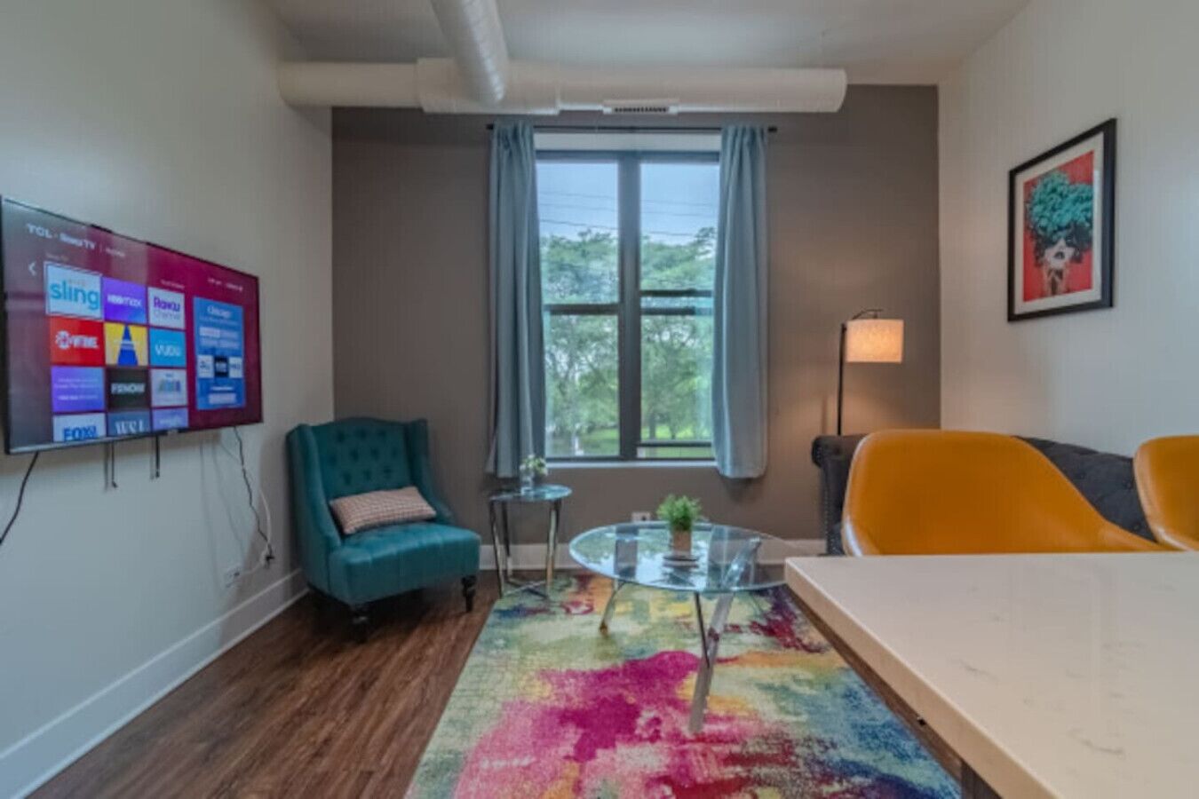 A modern room with a teal armchair, a yellow armchair, a glass coffee table, and a colorful rug. A flat-screen TV is mounted on the wall, and a window with curtains is visible.